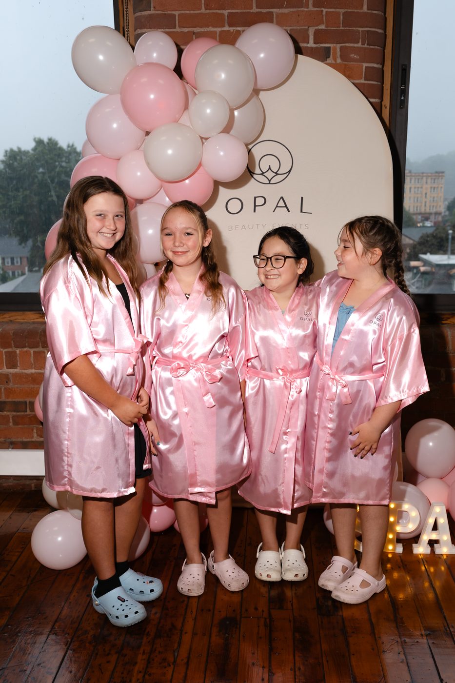 Four girls in pink spa robes laughing together in front of the Opal Beauty Events balloon backdrop