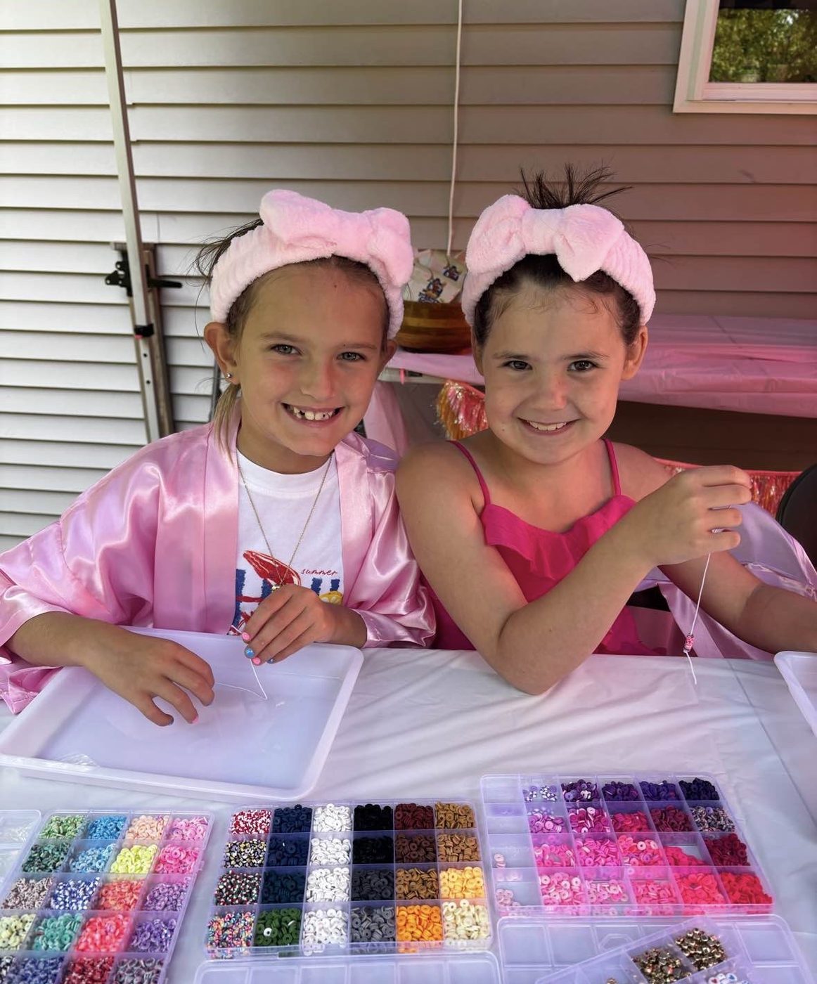 Two girls in pink robes and terry headbands smiling while picking out beads at the friendship bracelet station